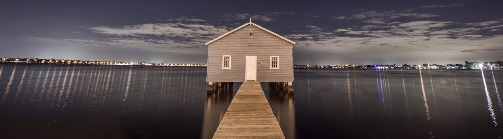 Blaues Bootshaus am Ende eines Holzstegs bei Nacht mit Lichtern am Horizont